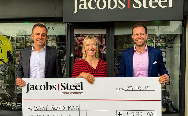 Two men and a woman smile and hold a giant cheque in front of estate agent window