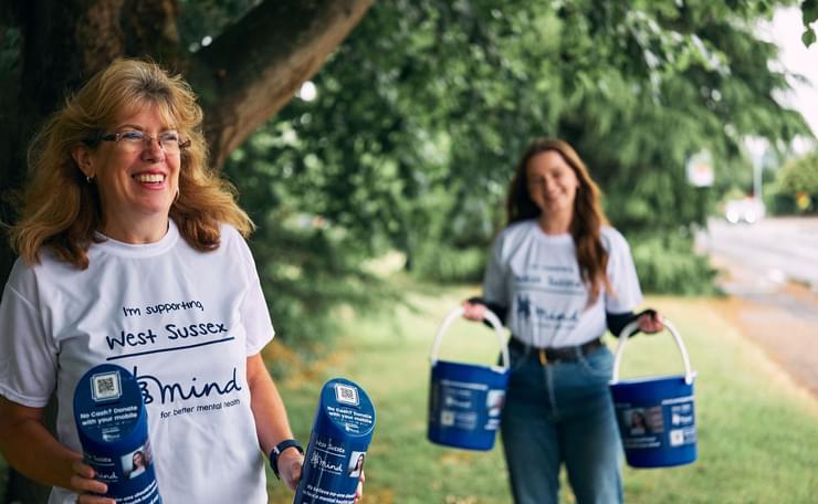 Two women in supporters' T-shirts hold collection tins and buckets with trees behind