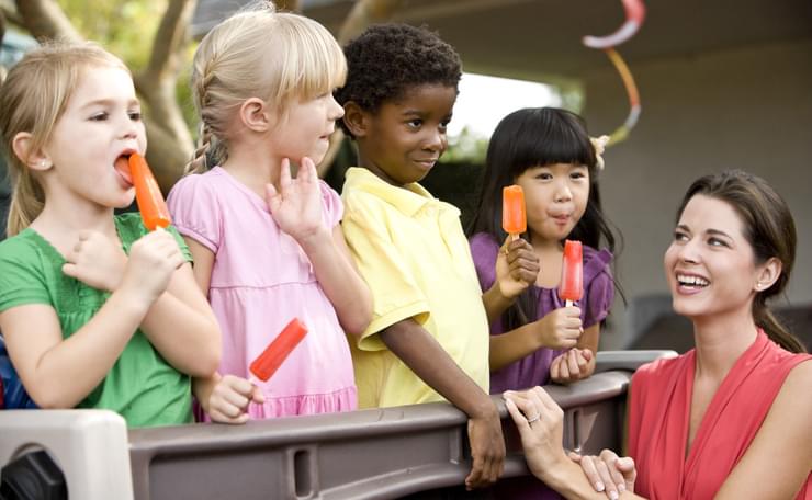 Four pre-school children eat ice lollies with young woman, at outside event