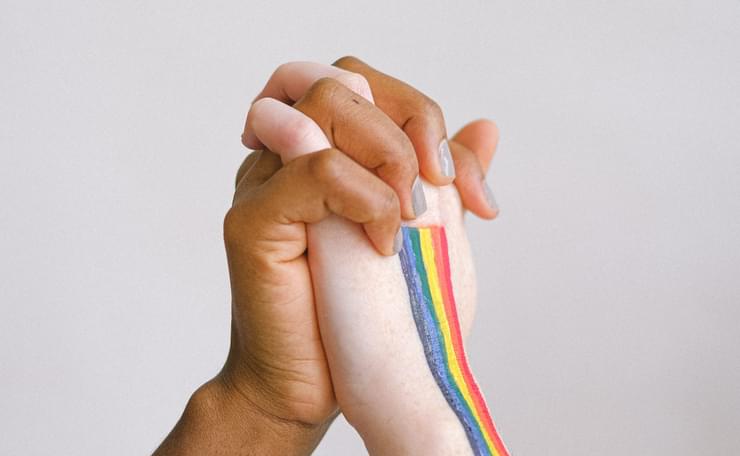 Picture of two hands holding, one black, one white, with rainbow flag on white hand