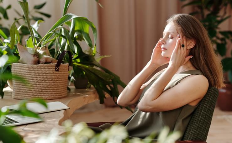 A woman gently rubs her temples to de-stress during Stress Awareness Month while sitting at a desk, with green plants around her