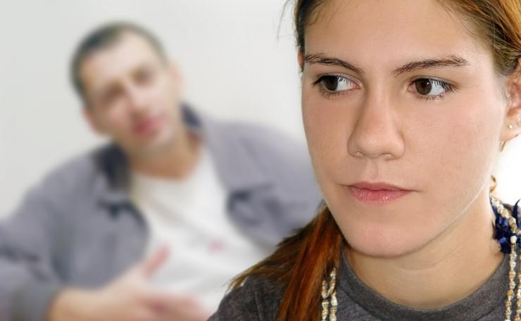 Unhappy teenage girl in foreground with concerned dad talking in background