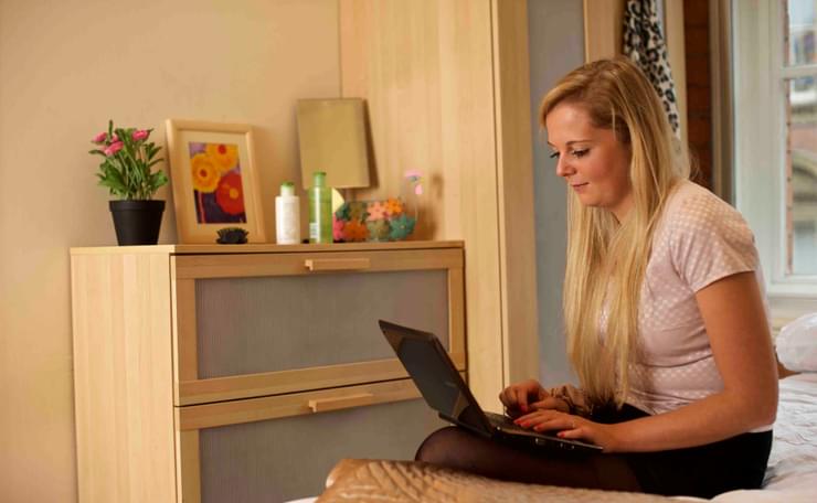 Woman looks at laptop whilst sitting on her bed in bedroom