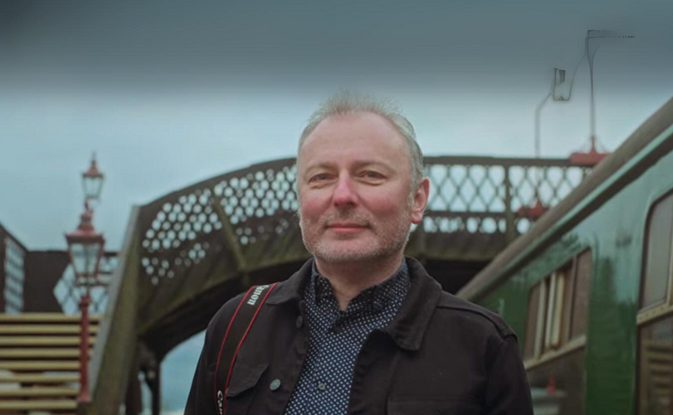 Man with grey hair in front of a bridge at a railway station with the train to his right