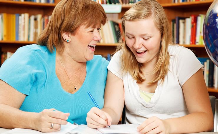 Happy woman and teenage girl at table in library with notebooks and pens