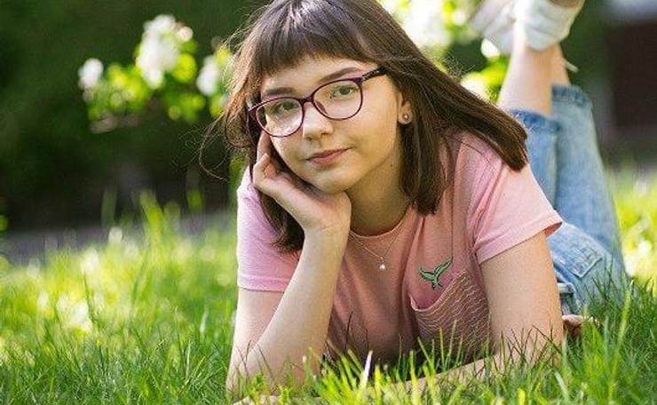 Young girl wearing pink T-shirt and glasses laying on grass with head rested in palm of hand